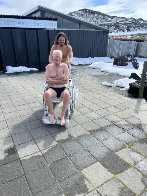 Guide pushing an older guest in a pool wheelchair across the paving at a geothermal pool, snow on the ground behind them