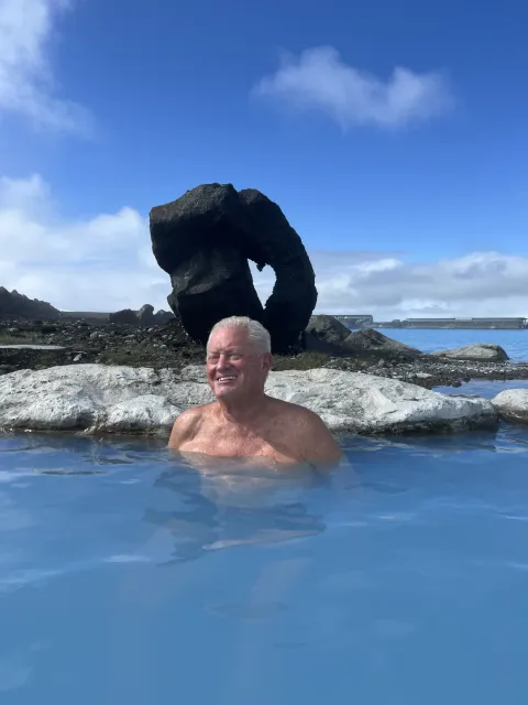 Older guest smiling shoulder-deep in a milky blue geothermal lagoon, a dark lava arch on the shore behind him