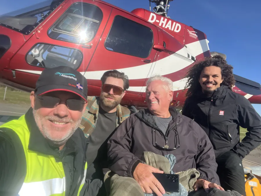 Guide and three travellers smiling in a selfie in front of a red Atlantsflug helicopter under a clear blue sky, one of them seated with a blanket over his legs