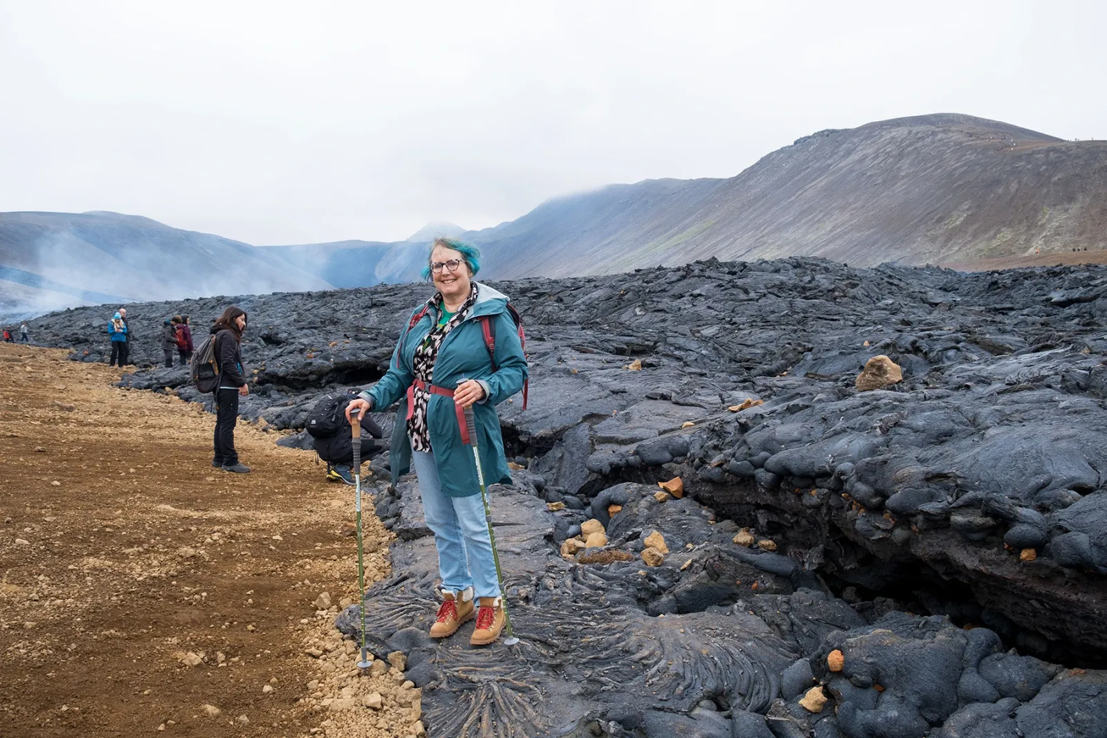 Traveller with walking poles standing on a cooled black lava field in Iceland, steam drifting from the hills behind her