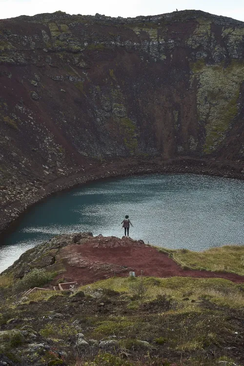 Traveller standing on a rock ledge above the turquoise water of Kerið crater lake, ringed by red volcanic walls on the Golden Circle