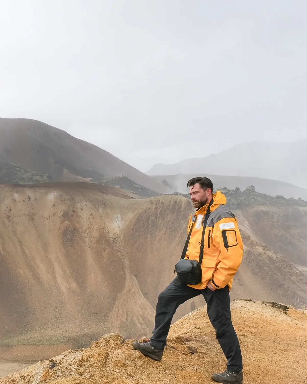Luke in an orange jacket standing on the rim of a rhyolite crater in the Icelandic Highlands, mist over the mountains behind him