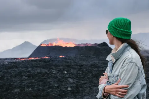 Traveller in a green beanie watching an erupting fissure on Reykjanes, glowing lava across the black lava field
