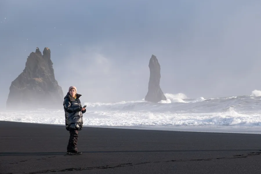 Traveller standing on the black sand of Reynisfjara, the Reynisdrangar sea stacks and breaking surf behind her