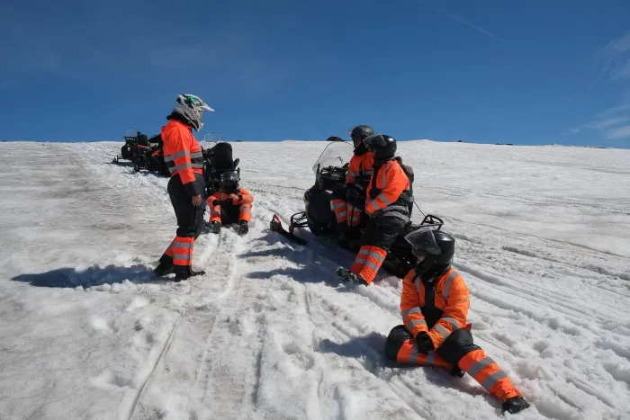 Group in orange snowsuits and helmets resting beside their snowmobiles on a sunlit glacier in Iceland