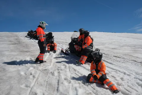 Group in orange snowsuits resting beside their snowmobiles on a sunlit glacier