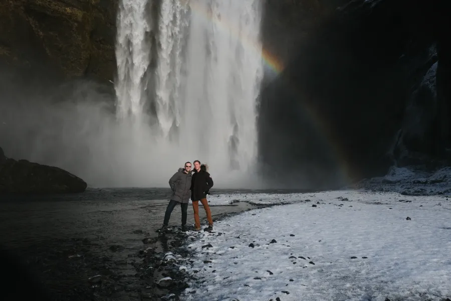 Couple standing on the snowy bank below Skógafoss in winter, a rainbow in the spray beside them