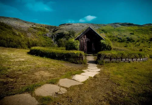 The reconstructed turf church at Þjóðveldisbærinn Stöng, a stone path leading to its wooden door through green Highland hills