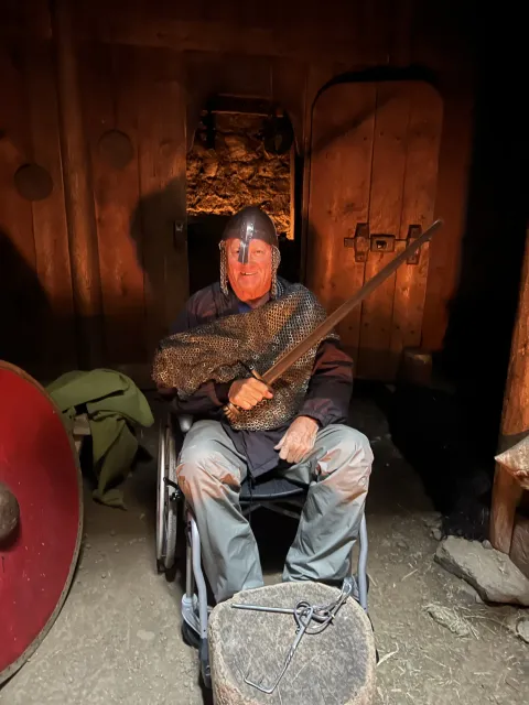 Guest in a wheelchair wearing a Viking helmet and chainmail, holding a sword inside a dim wooden turf house