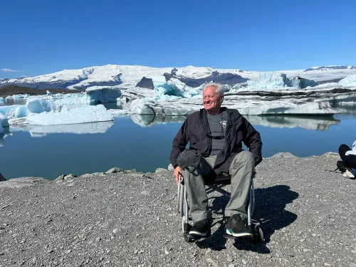 Smiling traveller in a wheelchair on the gravel shore of Jökulsárlón glacier lagoon, icebergs and the glacier behind him