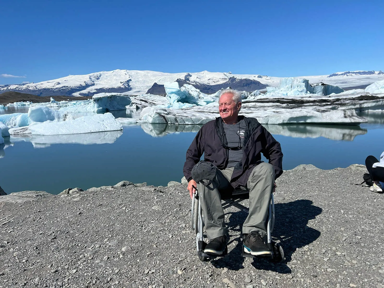 Smiling traveller in a wheelchair on the gravel shore of Jökulsárlón glacier lagoon, icebergs and the glacier behind him